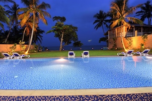 Piscina com espreguiçadeiras, palmeiras e vista para o oceano ao entardecer.