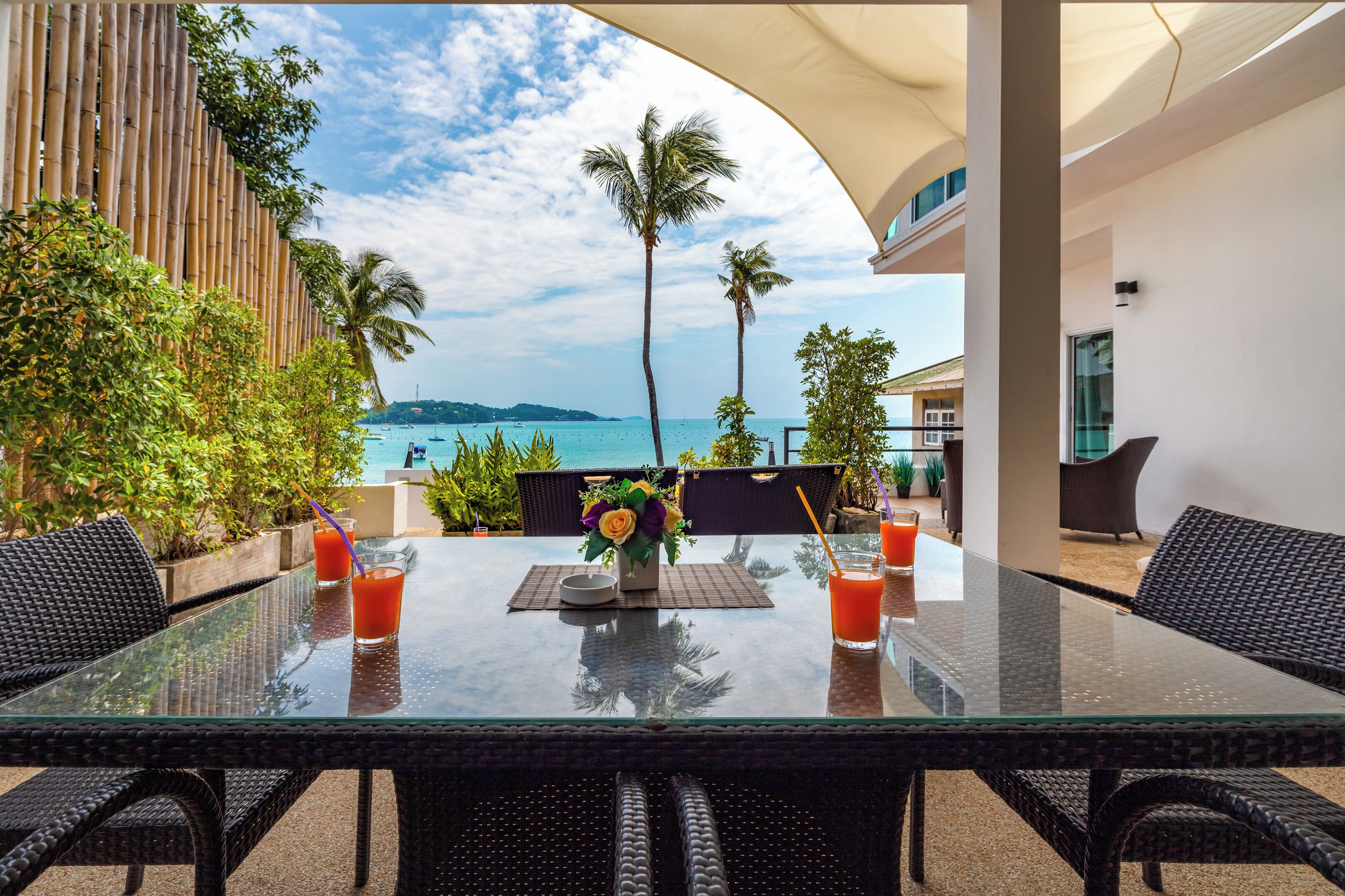 A glass-top table with two drinks, a vase with flowers, and a view of the sea and palm trees.