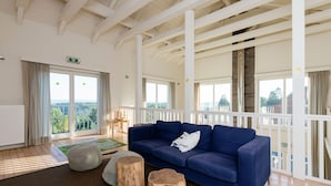 A spacious room with a blue sofa, wooden stools, and a view of the outdoors through large windows.