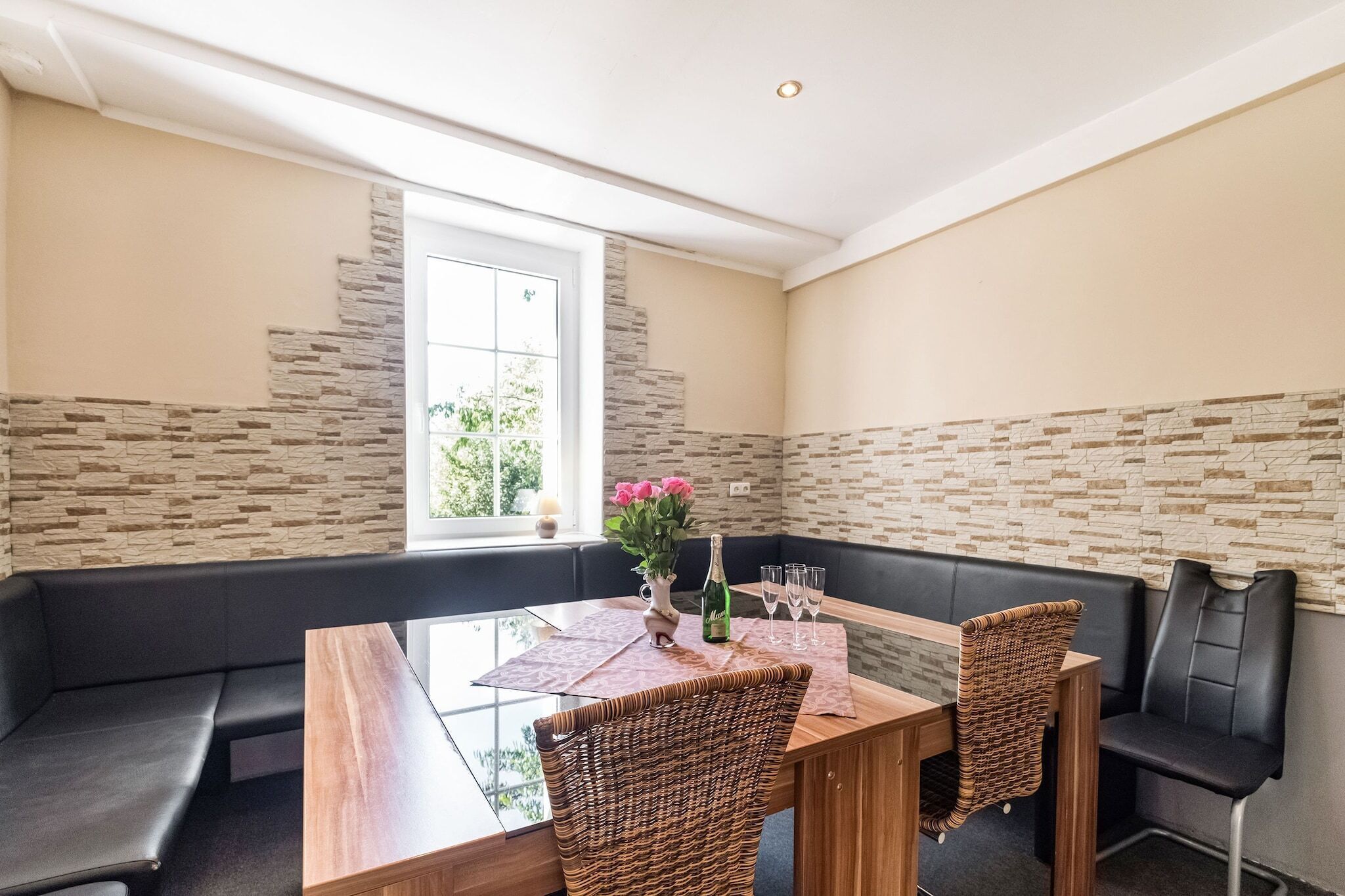 Een eethoek met een glazen tafel, een zwarte gewatteerde bank, rieten stoelen en een backsplash van natuursteen.