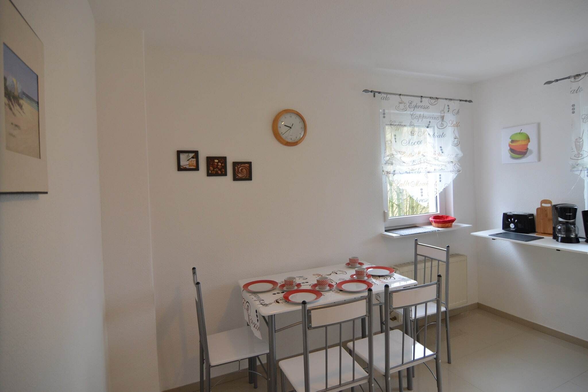 Ett litet kök med ett matbord för fyra personer, en klocka på väggen och ett fönster med gardiner.