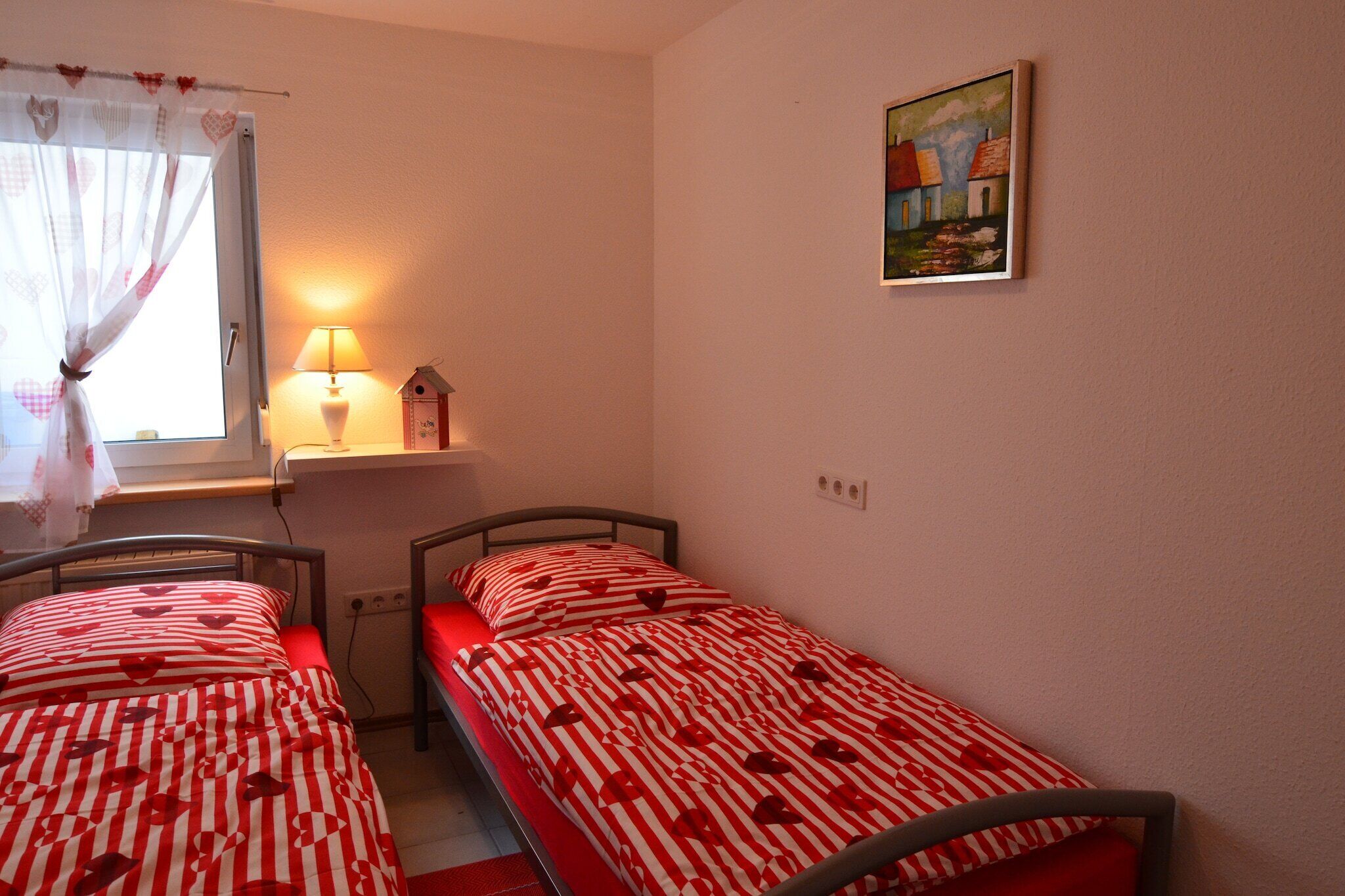 Un dormitorio con dos camas individuales, una ventana con cortina, una lámpara de noche y un cuadro en la pared.