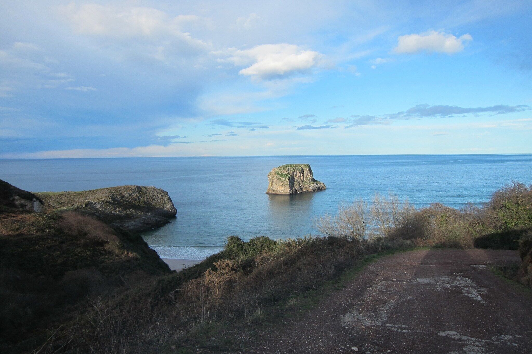Eine Küstenlandschaft mit einem Felsen im Meer, einem Feldweg, der zum Wasser führt, und einem wolkenlosen Himmel.