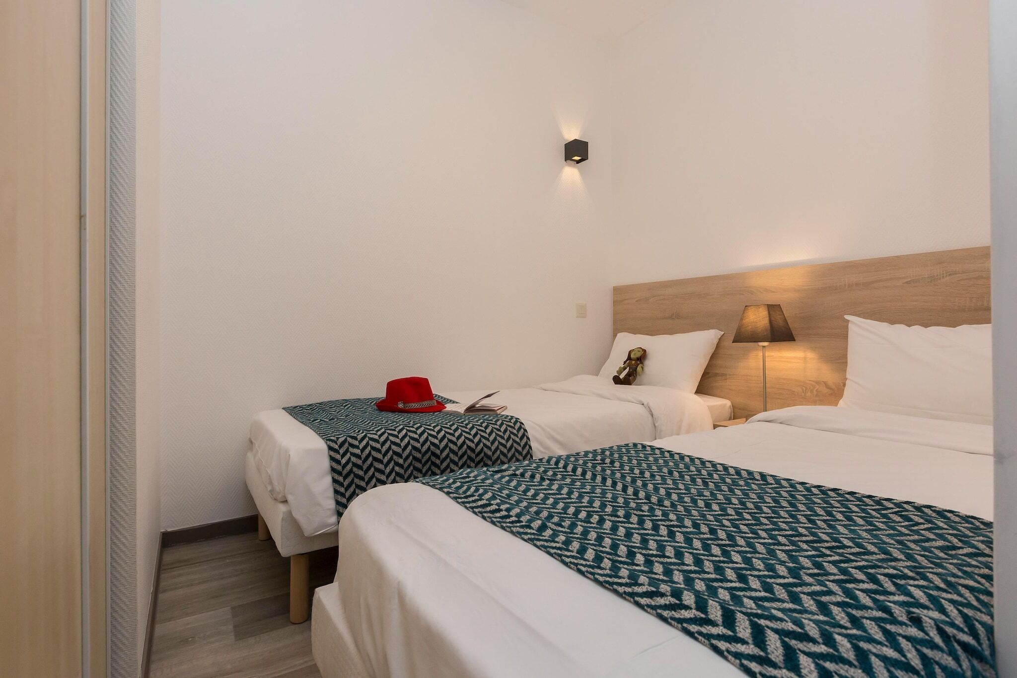 A hotel room with two beds, a nightstand, a lamp, and a wall-mounted light fixture.