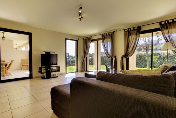 A living room with a large sofa, a TV, and a view of a garden through large windows.
