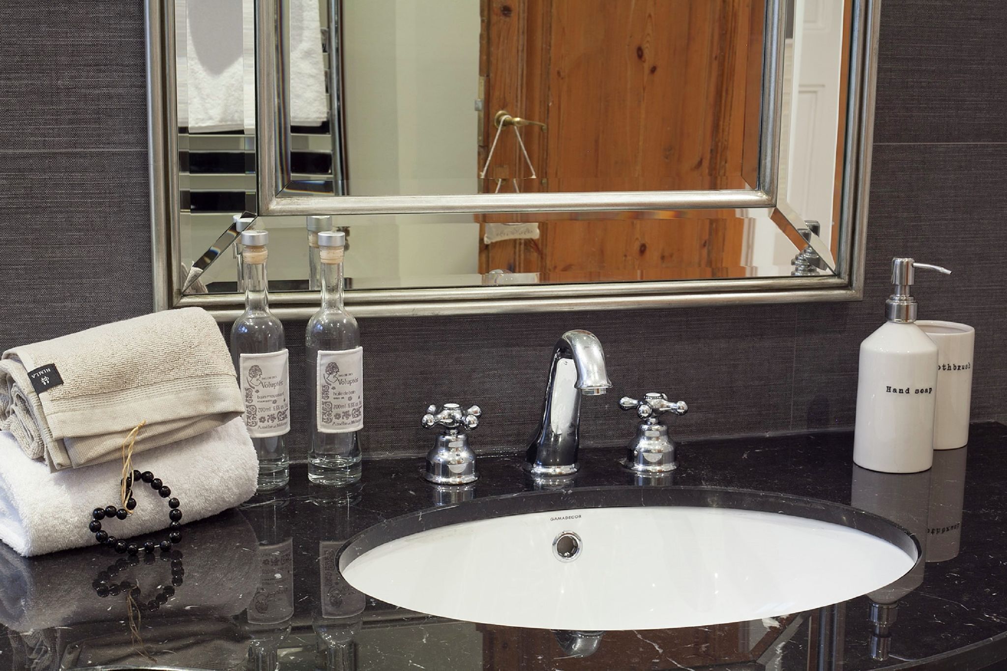 Un baño con un lavabo blanco, dos llaves, un espejo y artículos de aseo que incluyen una toalla, botellas y un dispensador de jabón.