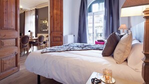 A hotel room with a large bed, a wooden wardrobe, a dining table with chairs, and a view of the city through the window.