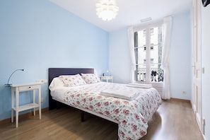 Een slaapkamer met een bed, nachtkastje, lamp en een raam met uitzicht op gebouwen.