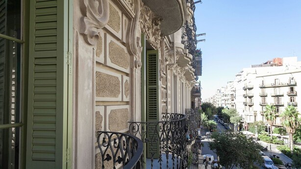 A balcony with ornate metal railings and green shutters, overlooking a street with parked cars and buildings.