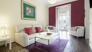 A living room with a white sofa, a coffee table, and a patterned rug. There is a painting on the wall and a door leading to another room.