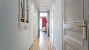 A hallway with a patterned floor, a white door with a brass handle, and a view into a room with a red wall and a bed.