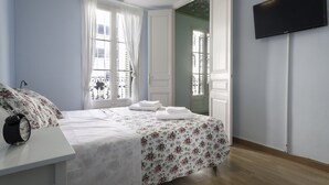 A bedroom with a bed, a bedside table with an alarm clock, a TV mounted on the wall, and a window with curtains.