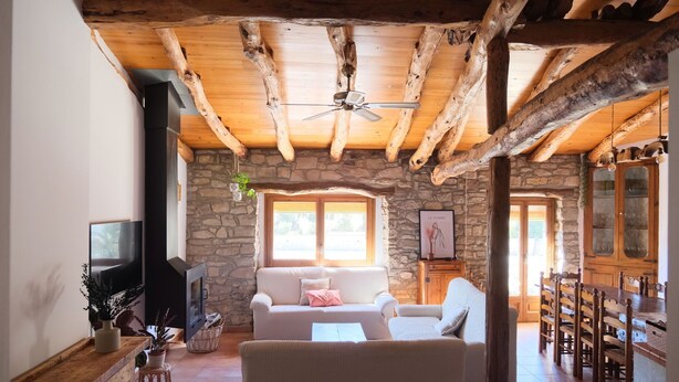 Un salon rustique avec des murs en pierre, des poutres en bois, une cheminée, un canapé, un téléviseur et un coin repas.
