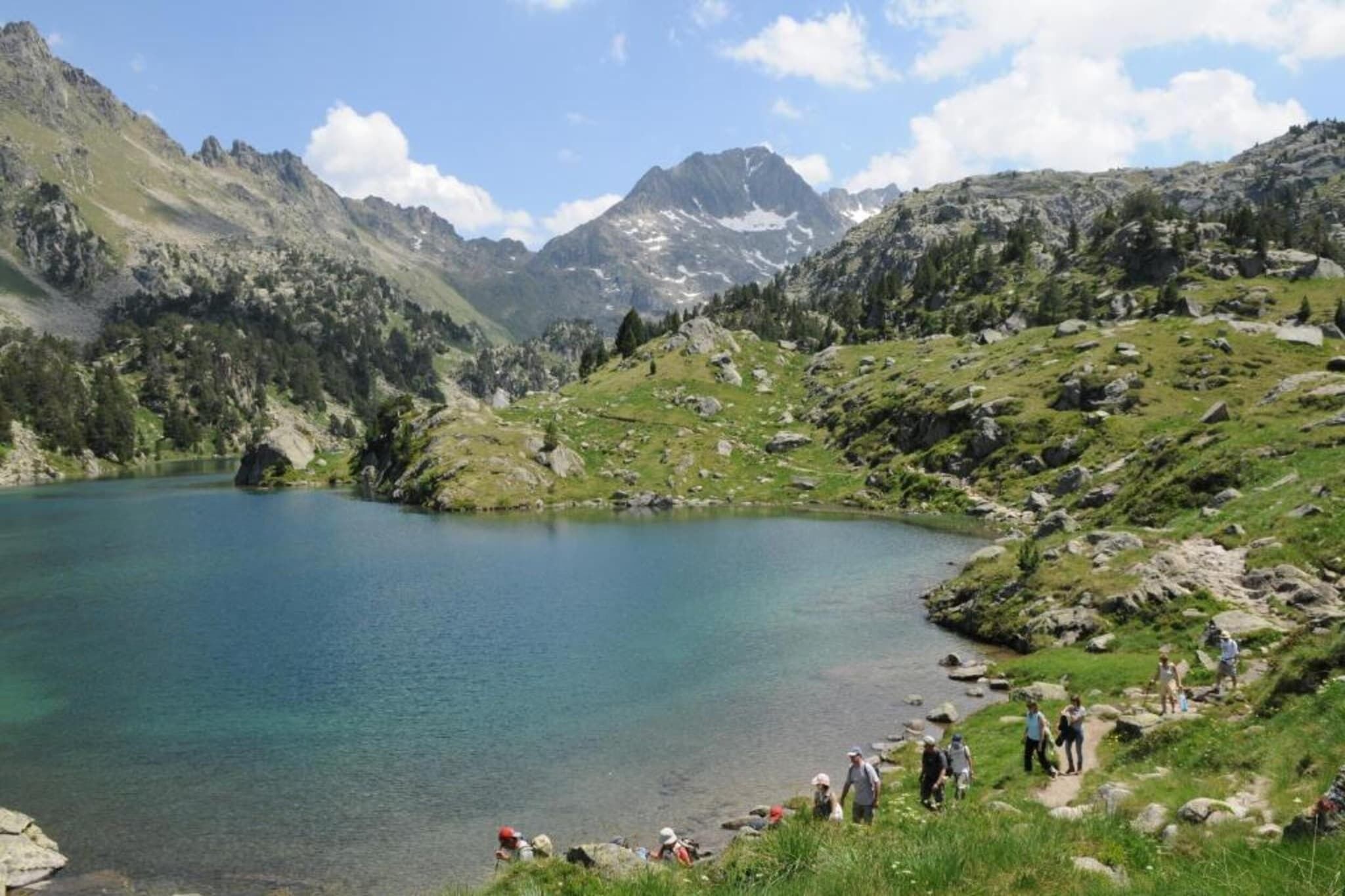A mountainous landscape with a clear lake, rocky terrain, and a group of people on a trail.