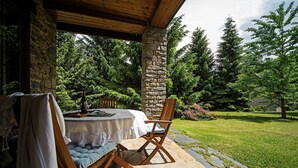 Un patio al aire libre con una mesa de piedra y sillas de madera, rodeado por un muro de piedra y abundante vegetación.
