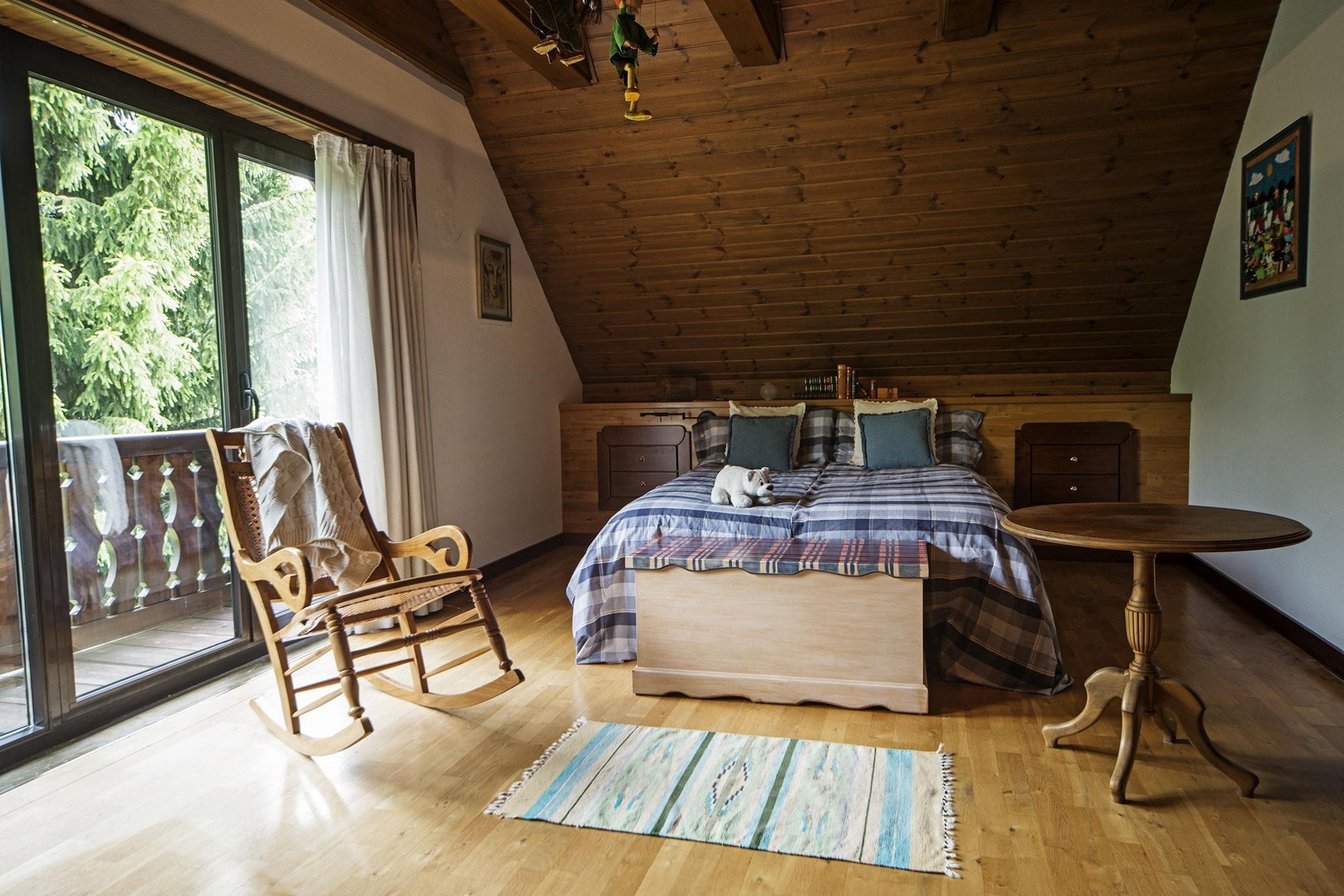 A bedroom with a wooden ceiling, a bed with a plaid bedspread, a rocking chair, a small round table, and a view of greenery outside.