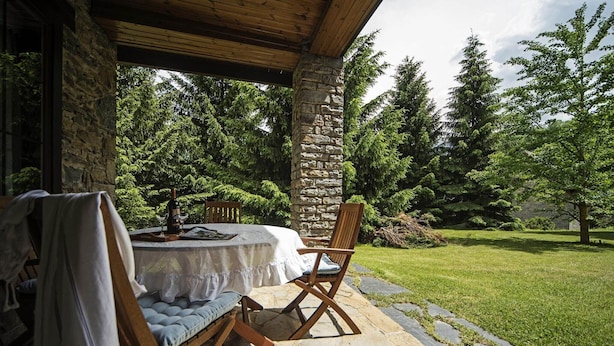 An outdoor patio with a stone table and wooden chairs, surrounded by a stone wall and lush greenery.