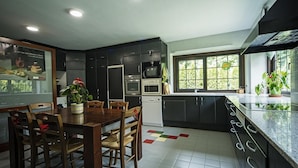 A modern kitchen with dark cabinets, a dining table with chairs, a refrigerator, an oven, a microwave, a dishwasher, and a window with a view of greenery.