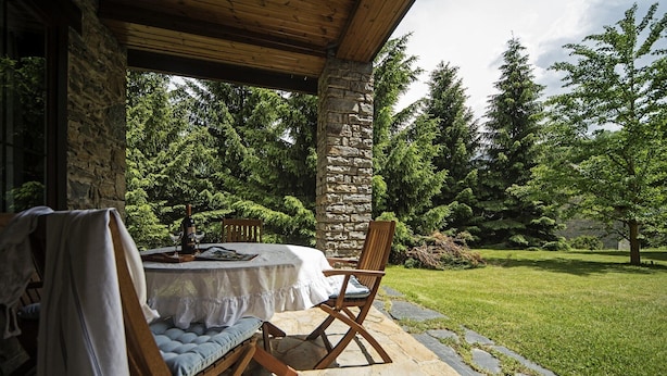 Un patio al aire libre con una mesa de piedra y sillas de madera, rodeado por un muro de piedra y abundante vegetación.
