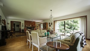 Amplia sala de estar con sofá, sillones, una mesa de centro y un comedor con mesa y sillas. Una ventana grande ofrece vistas al follaje exterior.