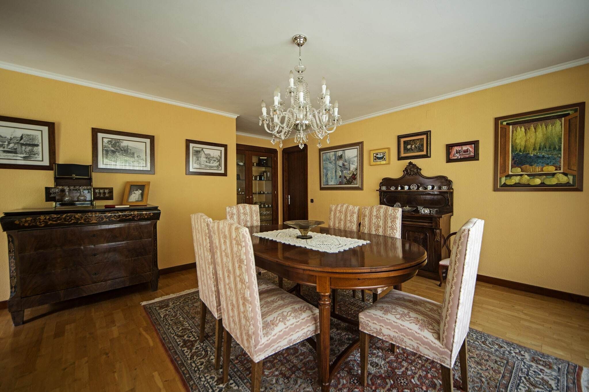 A dining room with a round table, chairs, a chandelier, framed pictures, and a wooden cabinet.