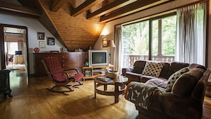 Una sala de estar acogedora con pisos de madera, un sillón mecedor, un sofá, un televisor y una ventana con cortinas.