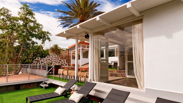 Uma área de lounge moderna ao ar livre, com espreguiçadeiras, um terraço envidraçado e vista para um edifício com fachada de pedra.