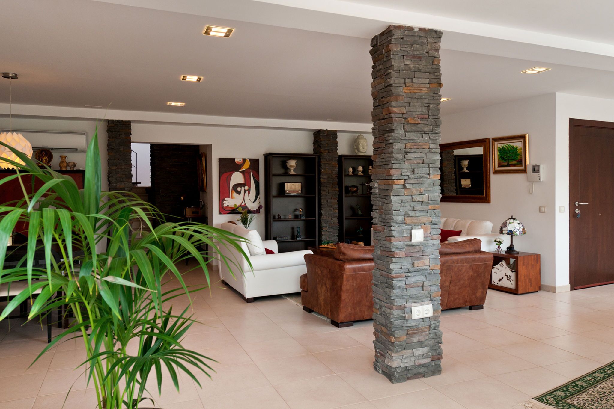 A spacious living area with a white sofa, a brown leather armchair, and a stone pillar. There are shelves with decorative items and a painting on the wall.