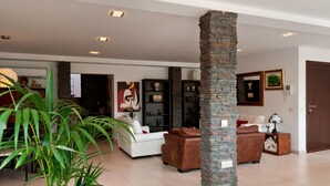 Uma sala de estar espaçosa com um sofá branco, uma poltrona de couro marrom e um pilar de pedra. Há prateleiras com objetos decorativos e um quadro na parede.