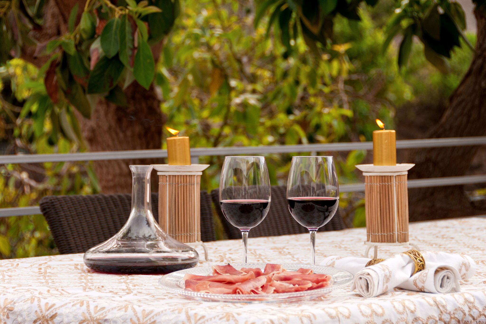 A table set with a glass decanter, two glasses of red wine, a plate of cured meats, and candles on a tablecloth with a natural backdrop.