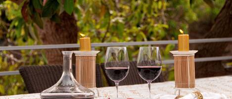 A table set with a glass decanter, two glasses of red wine, a plate of cured meats, and candles on a tablecloth with a natural backdrop.
