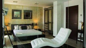 A modern bedroom with a large bed, a glass partition, a white armchair, and a wardrobe.