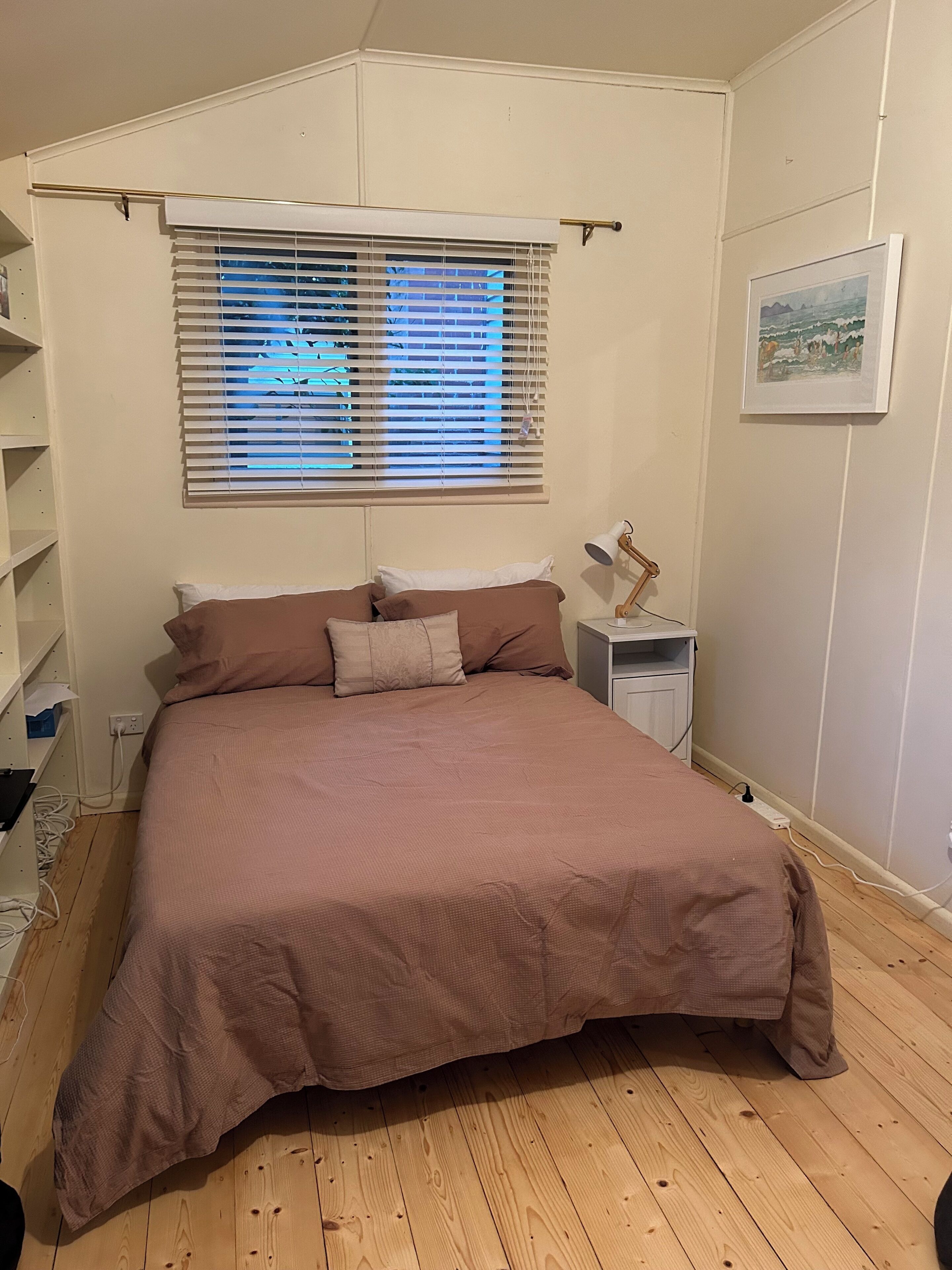 Un dormitorio con una cama grande, una mesita de noche con lámpara, una ventana con persianas y un cuadro enmarcado en la pared.