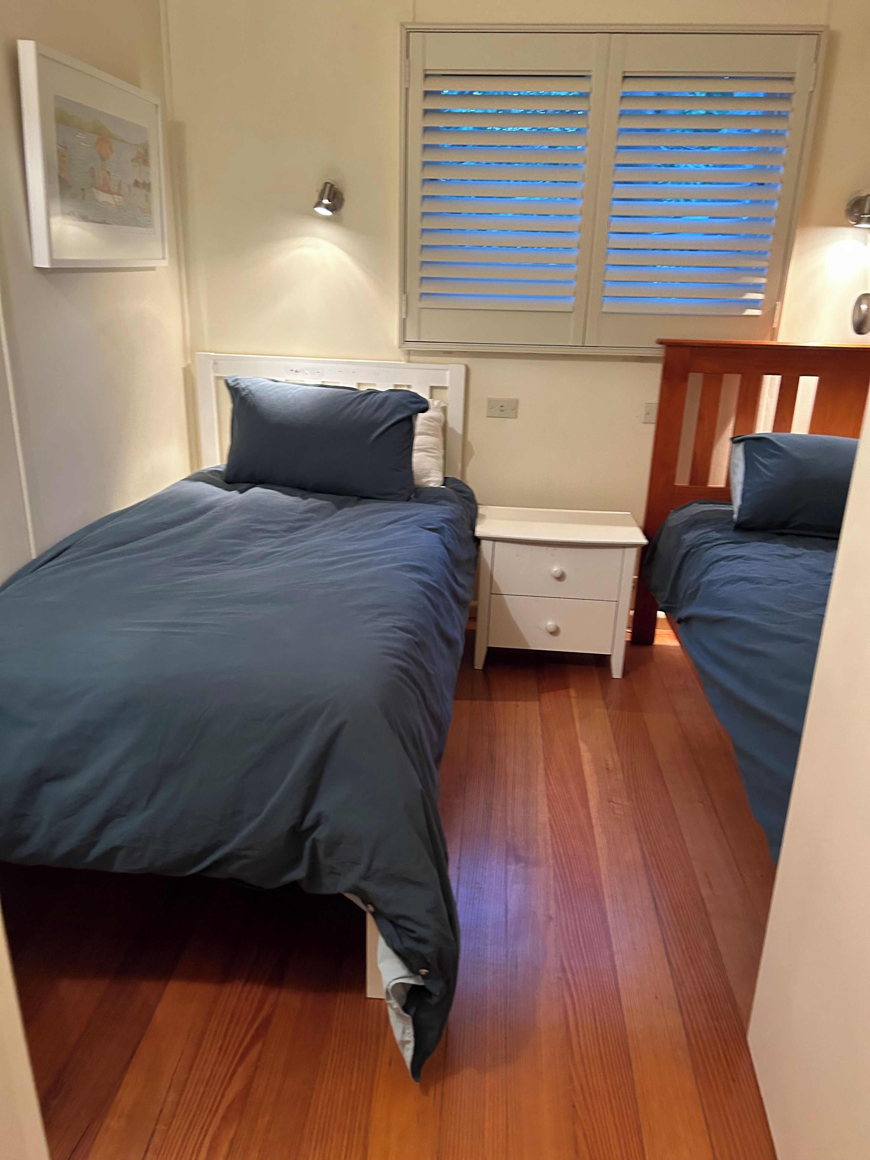 A small, compact room with two single beds, a nightstand, and a window with blinds.