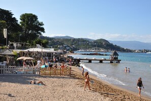 A beach with people sunbathing and walking, a pier extending into the water, and buildings in the background. - Villa in Var with Hill Views & Pool (Montauroux)