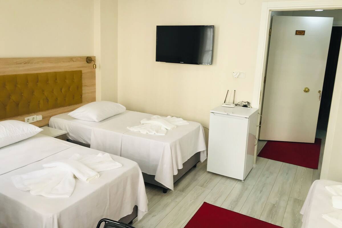A hotel room with two single beds, a TV mounted on the wall, a small desk with a printer, and a door with a number plate.