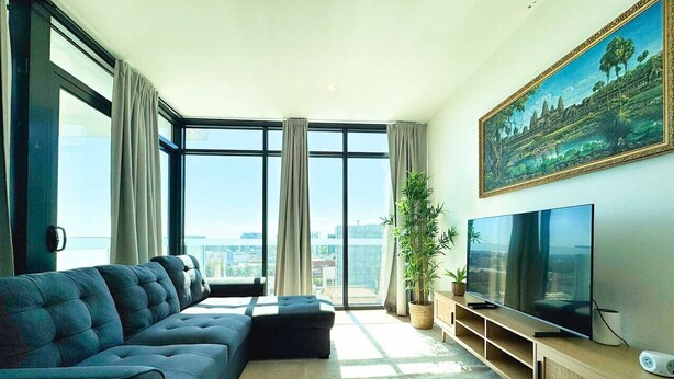 A modern living room with a large sofa, a flat-screen TV, and a city view through large windows. - Beautiful Cambodian Inspired Apt with Pool & Gym (Adelaide)