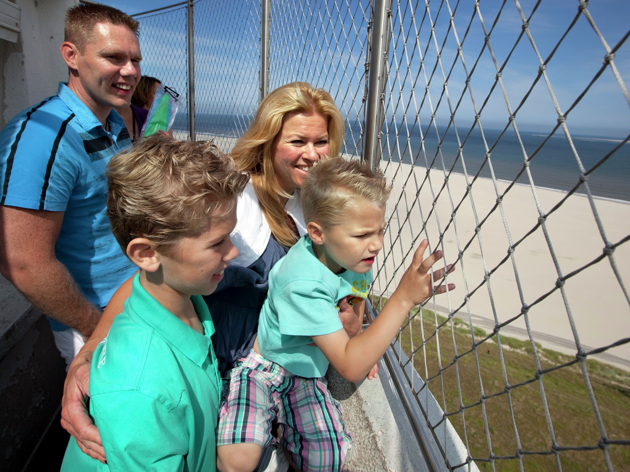 En familj på fyra personer befinner sig vid kusten, med ett staket av metall och utsikt över havet.