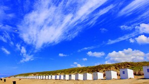 A row of white beach huts with doors, a sandy beach, and a clear blue sky with clouds. - Thatched wellness villa near Texel Sea (De Koog)