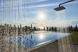 A swimming pool with a showerhead, surrounded by a paved area and trees, with a sunset in the background.