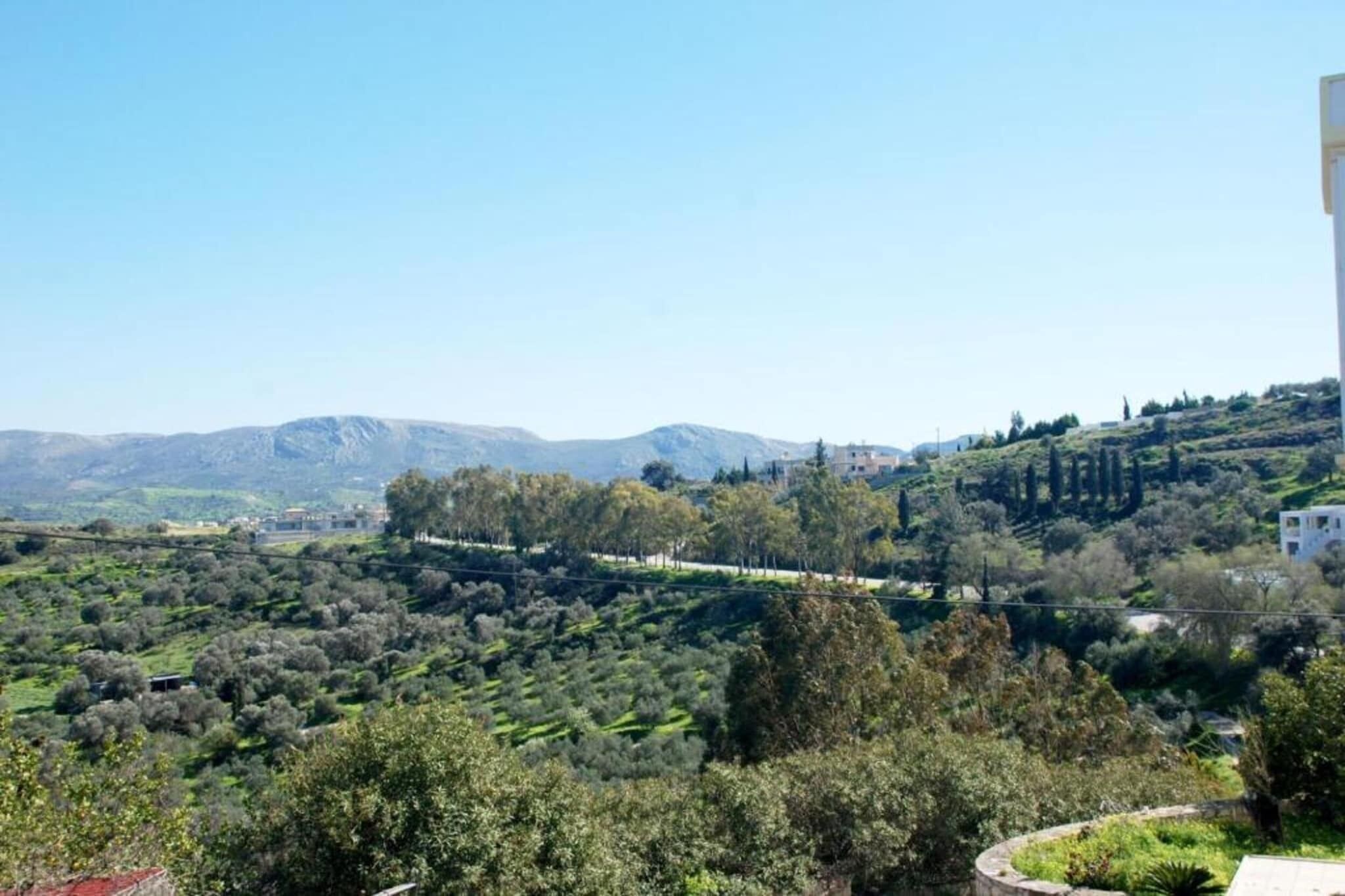 A hillside landscape with olive groves, a few buildings, and distant mountains under a clear sky.