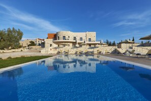 A swimming pool with lounge chairs and umbrellas, a stone building with balconies, and a clear blue sky. - Villa in Kamilari near Kid-Friendly Beach (Kamilari)