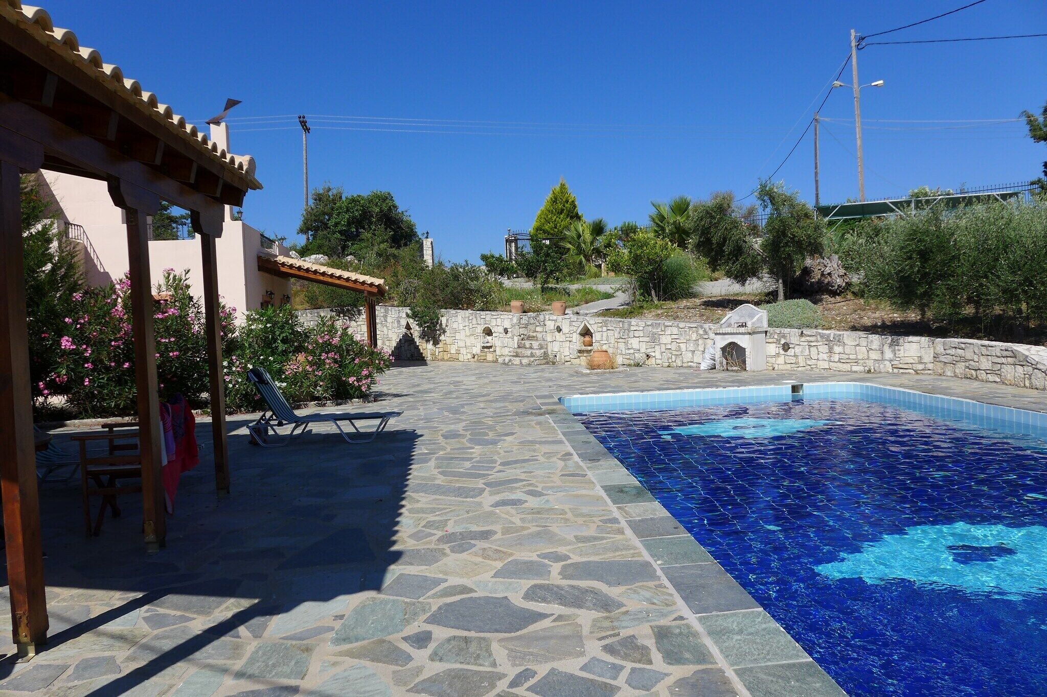 A swimming pool with stone flooring, a pergola, and a stone wall with a small arched entrance.