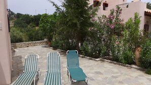 A patio with three striped lounge chairs and a stone floor, surrounded by greenery and a building with pink walls. - Villa in Axos with Pool near Panormo Beach (Axos - Rethymno)