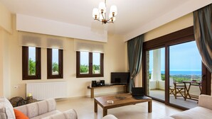 Un salon moderne avec un canapé, une table basse et un téléviseur. De grandes fenêtres sont agrémentées de rideaux, un balcon avec une table offre une vue sur la mer.