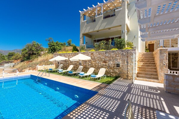 Un área de piscina con sillas de descanso, un muro de piedra y una escalera que lleva a un edificio con balcones.