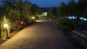 Un sendero de grava conduce a una casa con techo de tejas rojas, rodeada de vegetación e iluminada por luces de jardín.