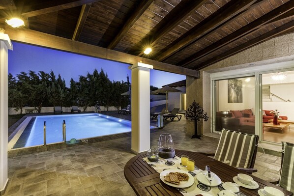 An outdoor pool area with a dining table set for breakfast, a lounge chair, and a view of the pool.