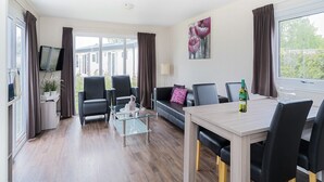 A modern living room with a glass coffee table, a sofa, and a dining table with chairs. - Chalet in Parc de Kievit with Private Garden (Baarle-Nassau)