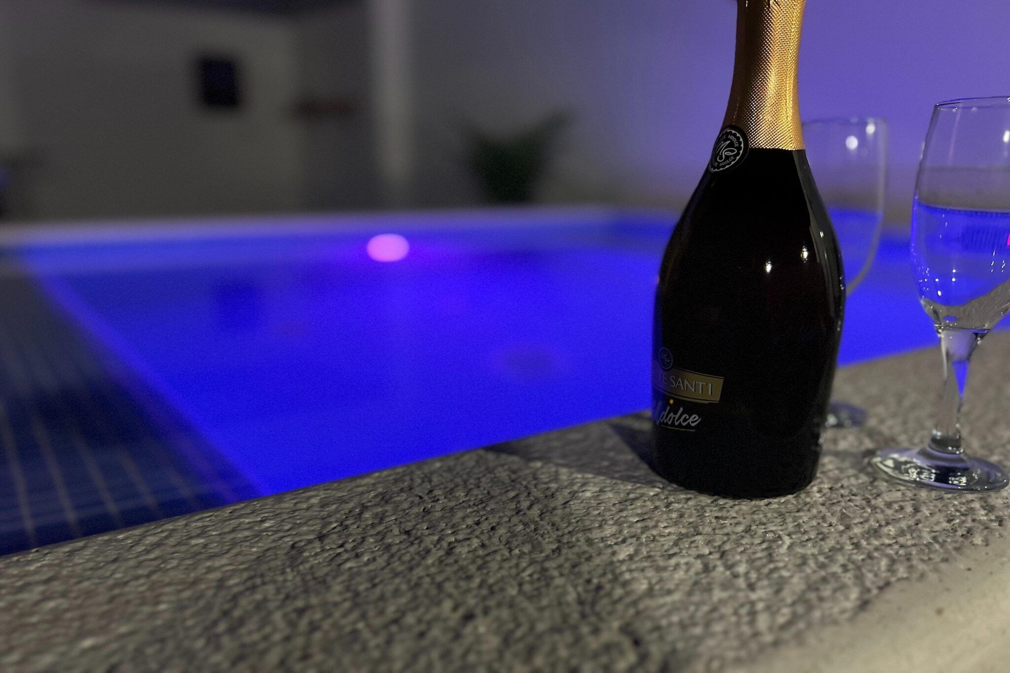 A bottle of Santi wine next to a glass on a stone surface by a pool with blue lighting.